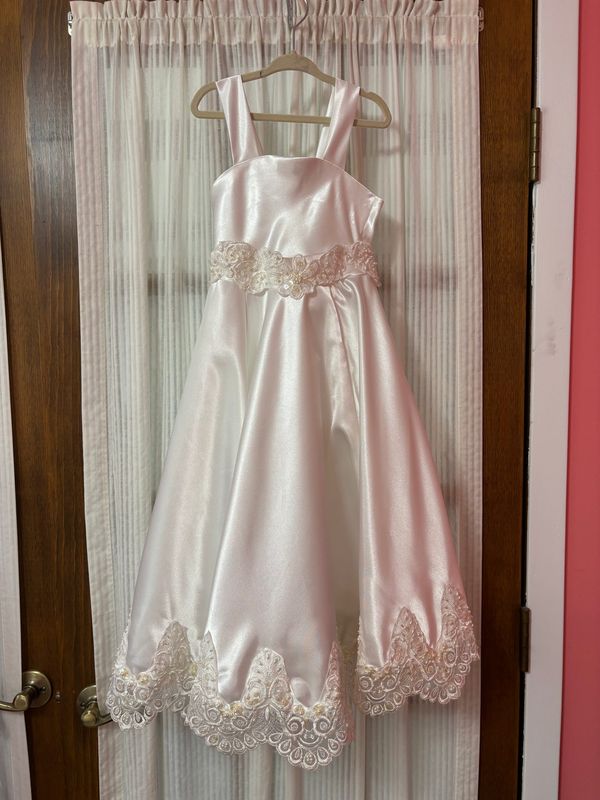 An ivory satin child’s dress with a lace hem on a hanger